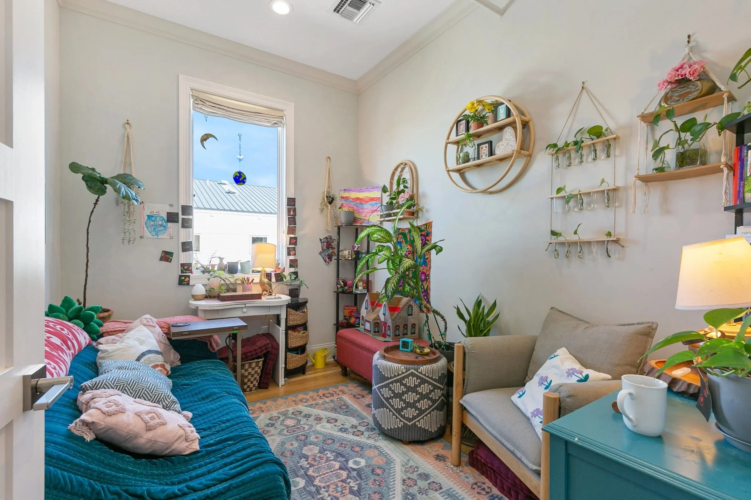 Cozy living room with a blue velvet sofa, beige armchair, and various plants and decorations; a window with hanging moon and globe ornaments, and wall shelves with small plants and decor.