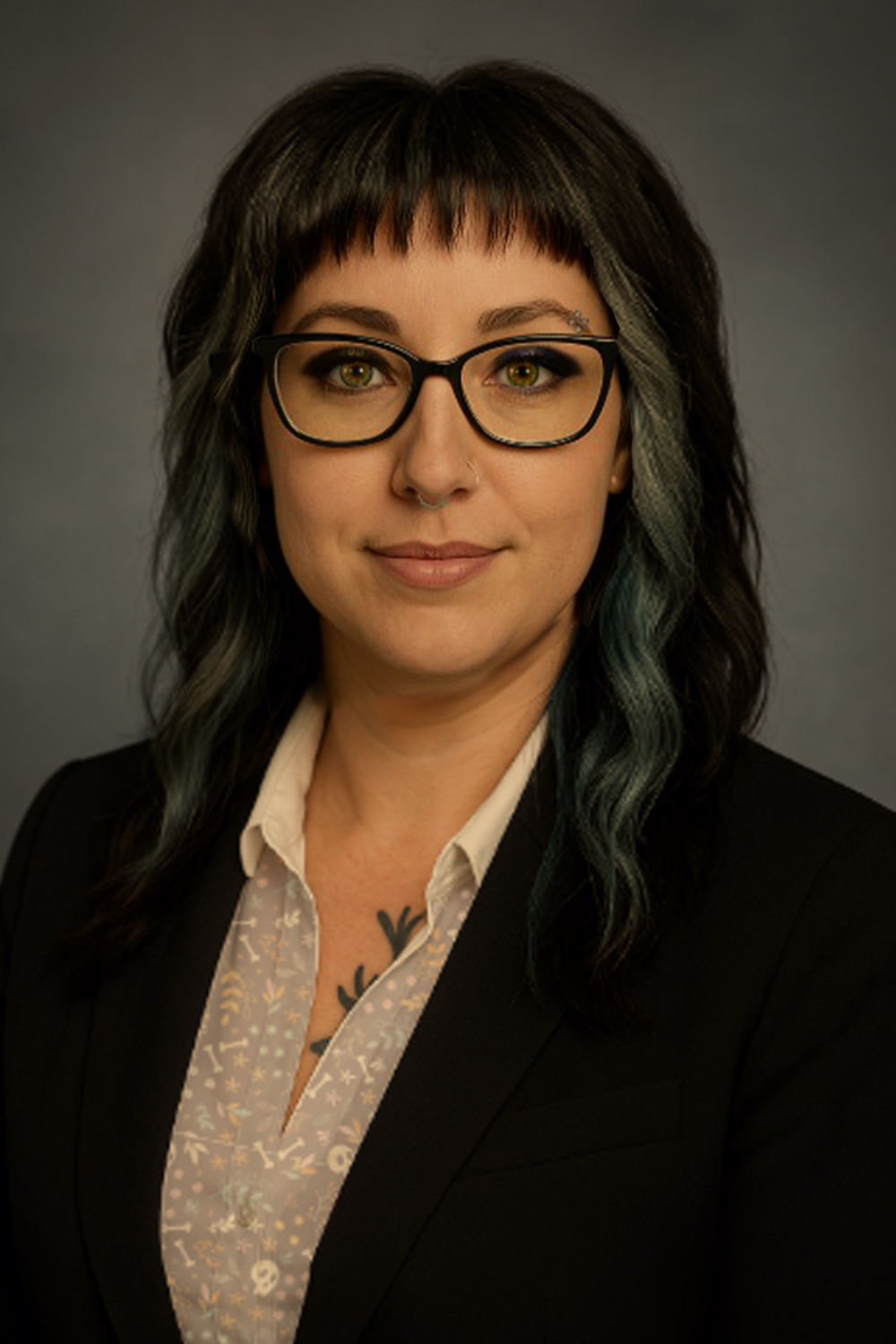 A woman with dark, shoulder-length hair with blue-green streaks, glasses, and a nose piercing, wearing a formal black blazer over a light patterned blouse, posing against a gray background.