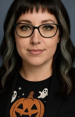A woman with black hair, glasses, and a septum piercing wearing a black shirt with a pumpkin design, posing against a plain background.