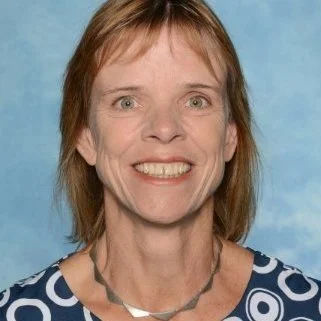 A woman with short, reddish-brown hair, smiling, wearing a blue and white patterned top and a silver necklace, against a blue background.