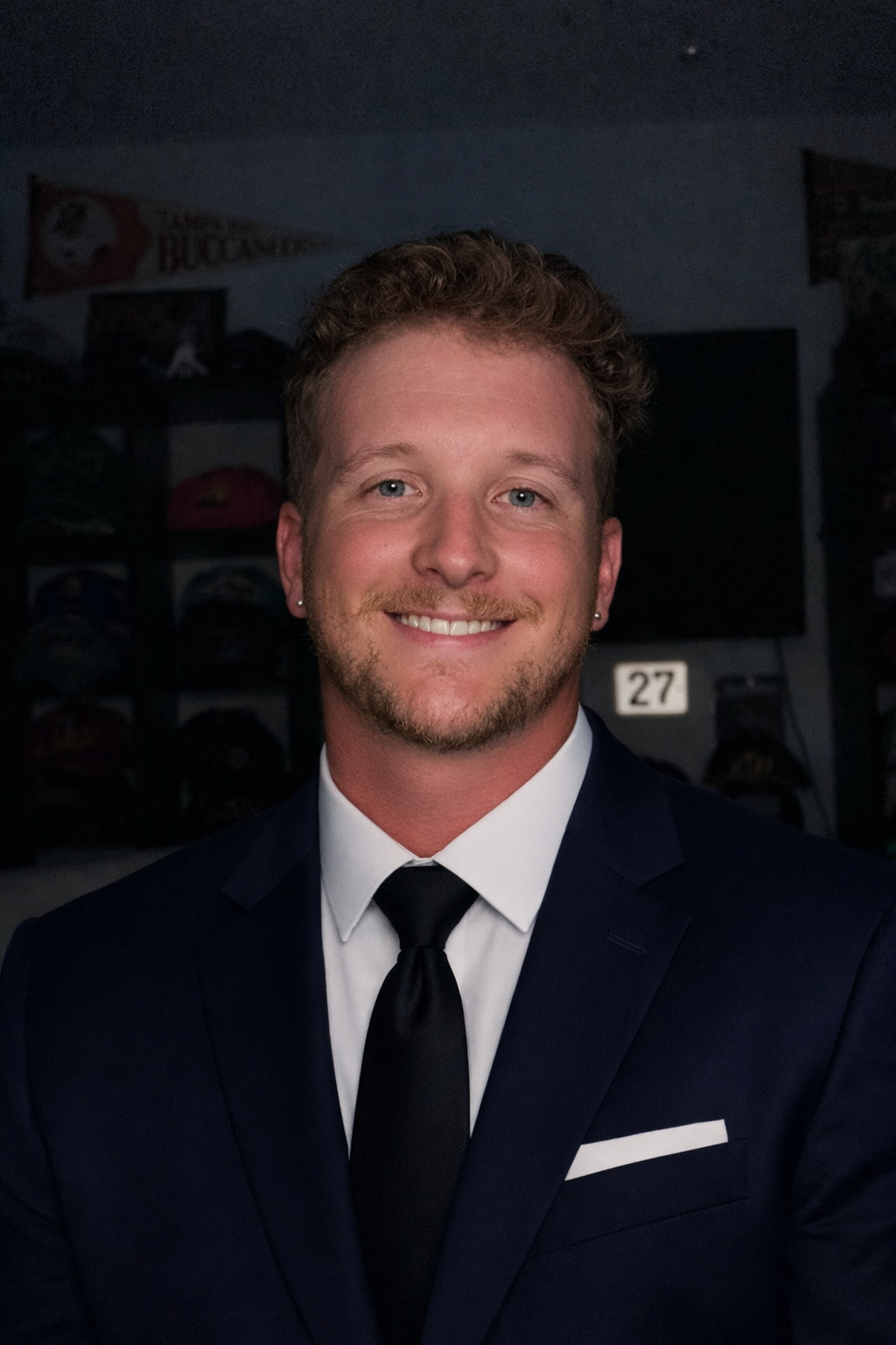 A young man in a dark suit, white shirt, and black tie smiling at the camera in a dimly lit room.
