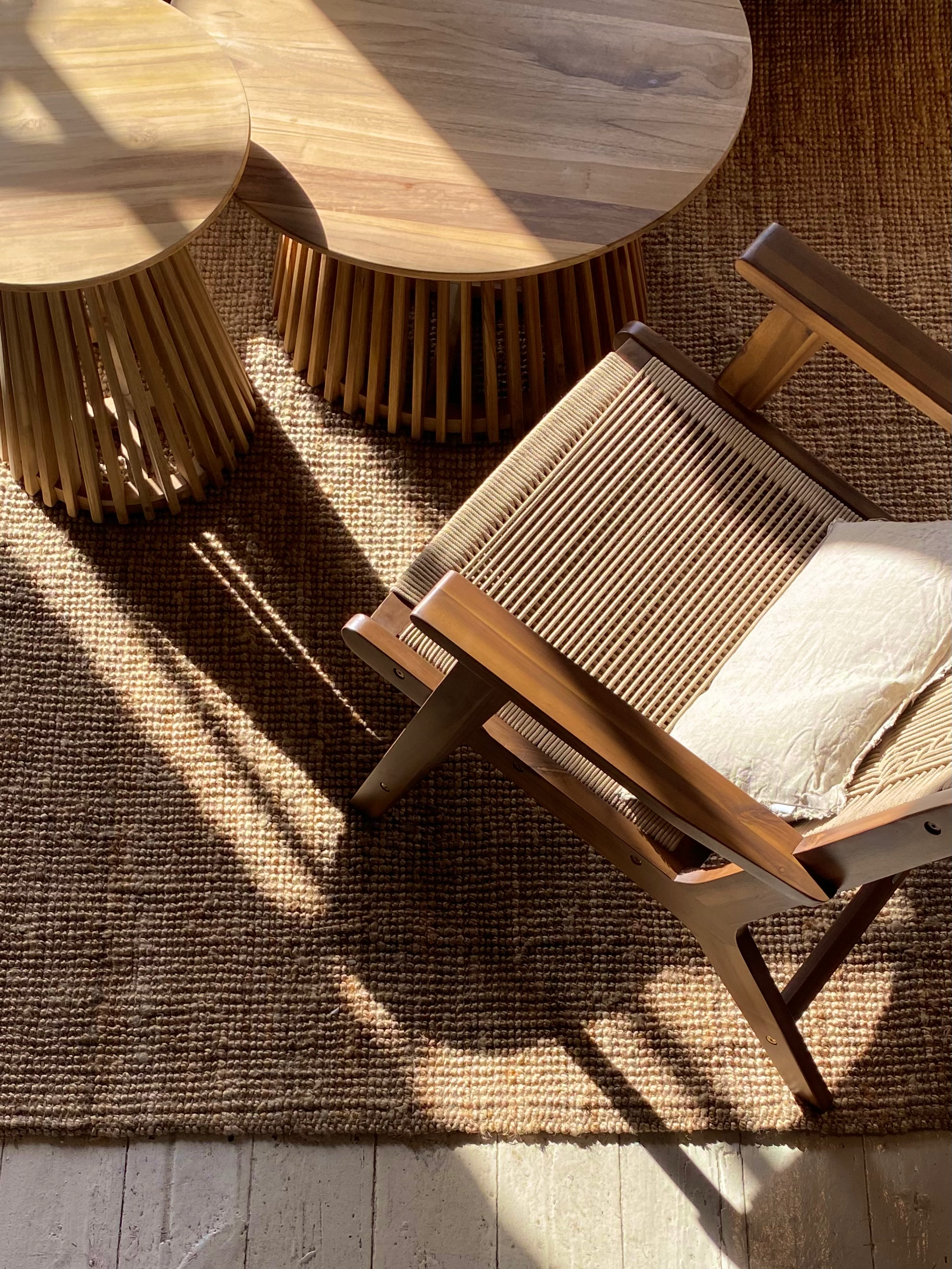 A wooden armchair with a light-colored cushion is placed on a textured woven rug. Two round wooden tables with slatted bases are beside the chair, with sunlight casting shadows across the scene.