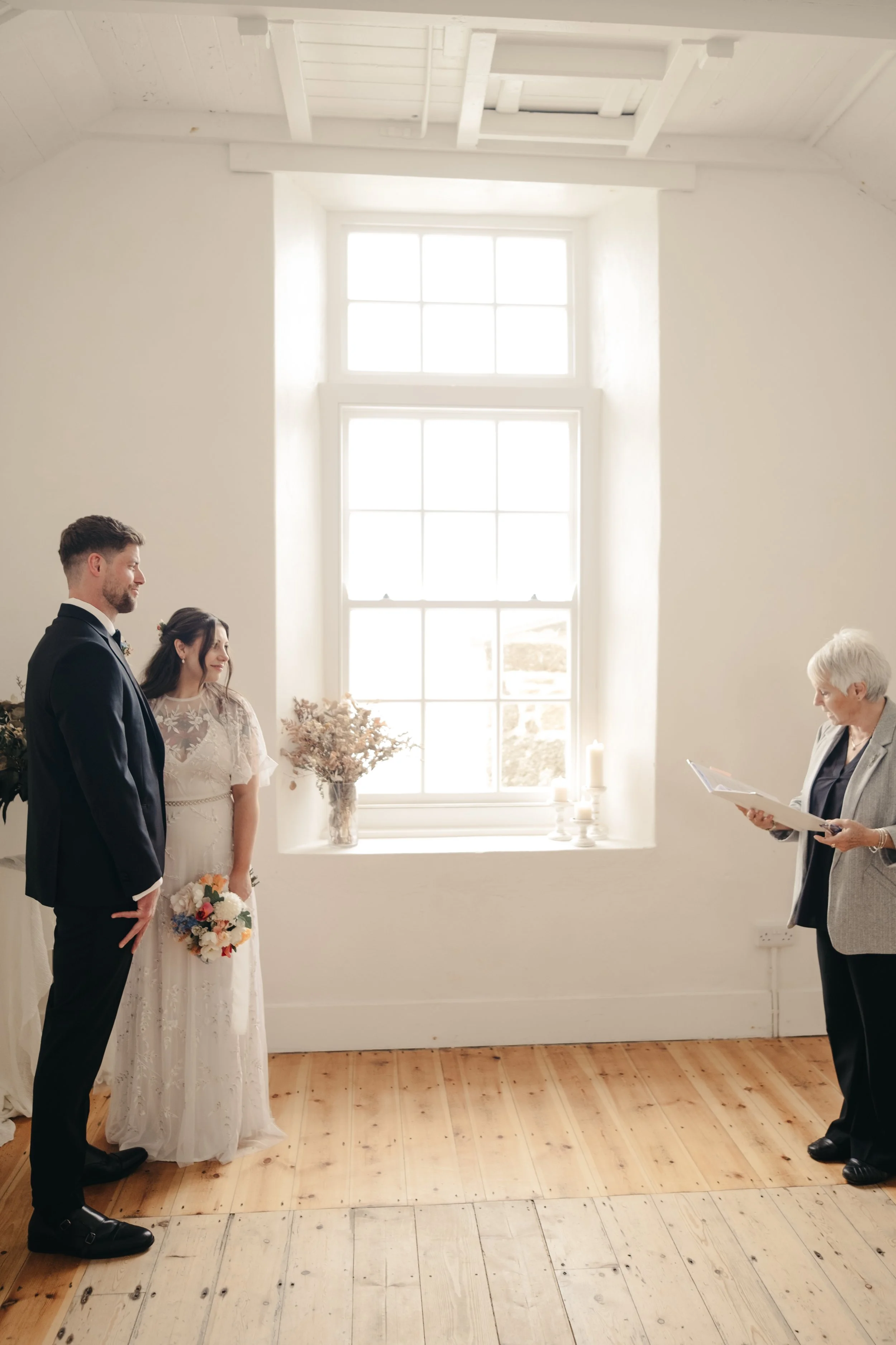 A wedding ceremony with a bride and groom standing in front of an officiant in a bright, white room with a large window and wooden floors.  St Ives, The Block House elopement venue, Cornwall.