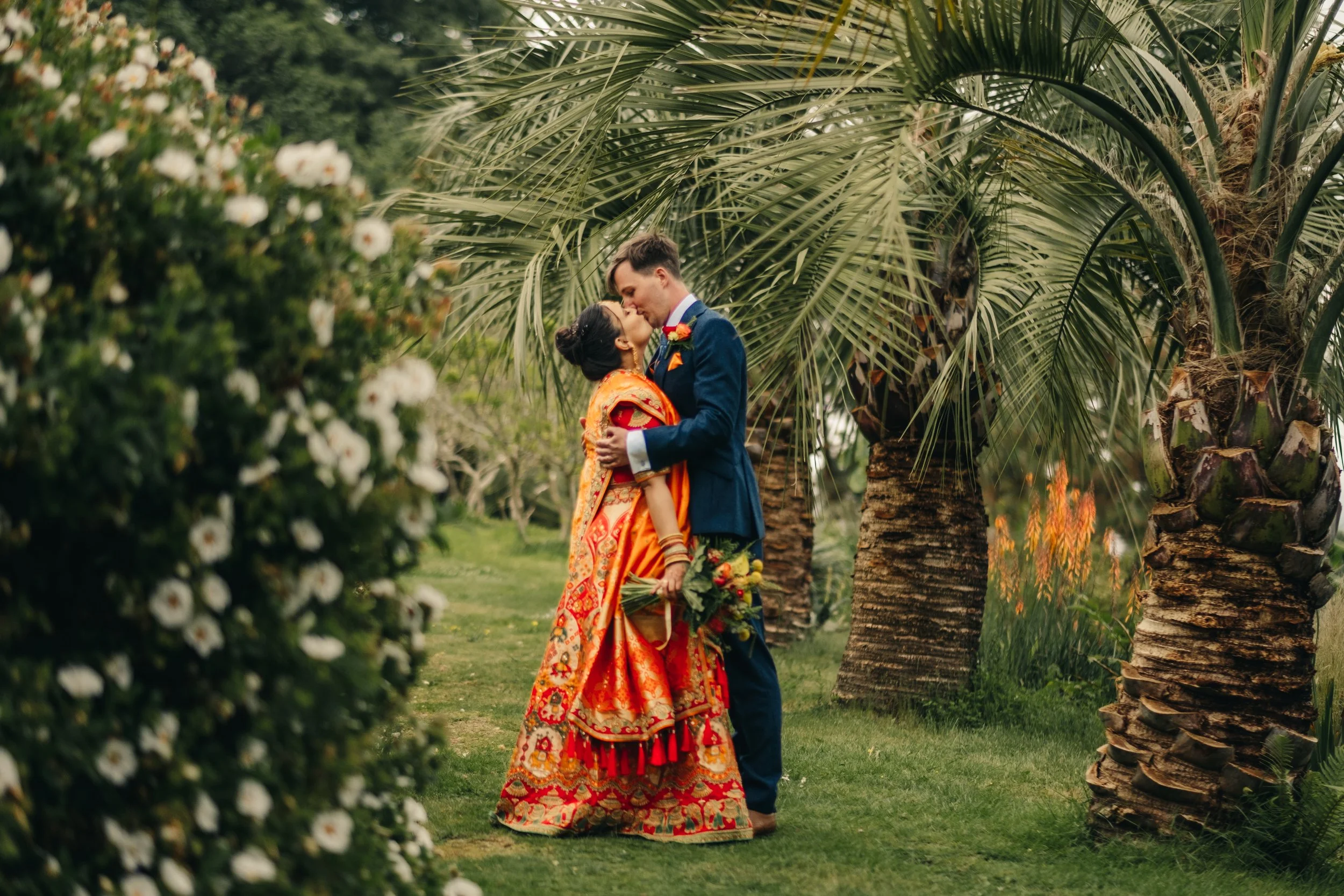 A couple in wedding attire sharing a kiss outdoors, surrounded by palm trees and blooming shrubs.Tremenheere Sculpture Gardens, Cornwall, Elopement Venue.