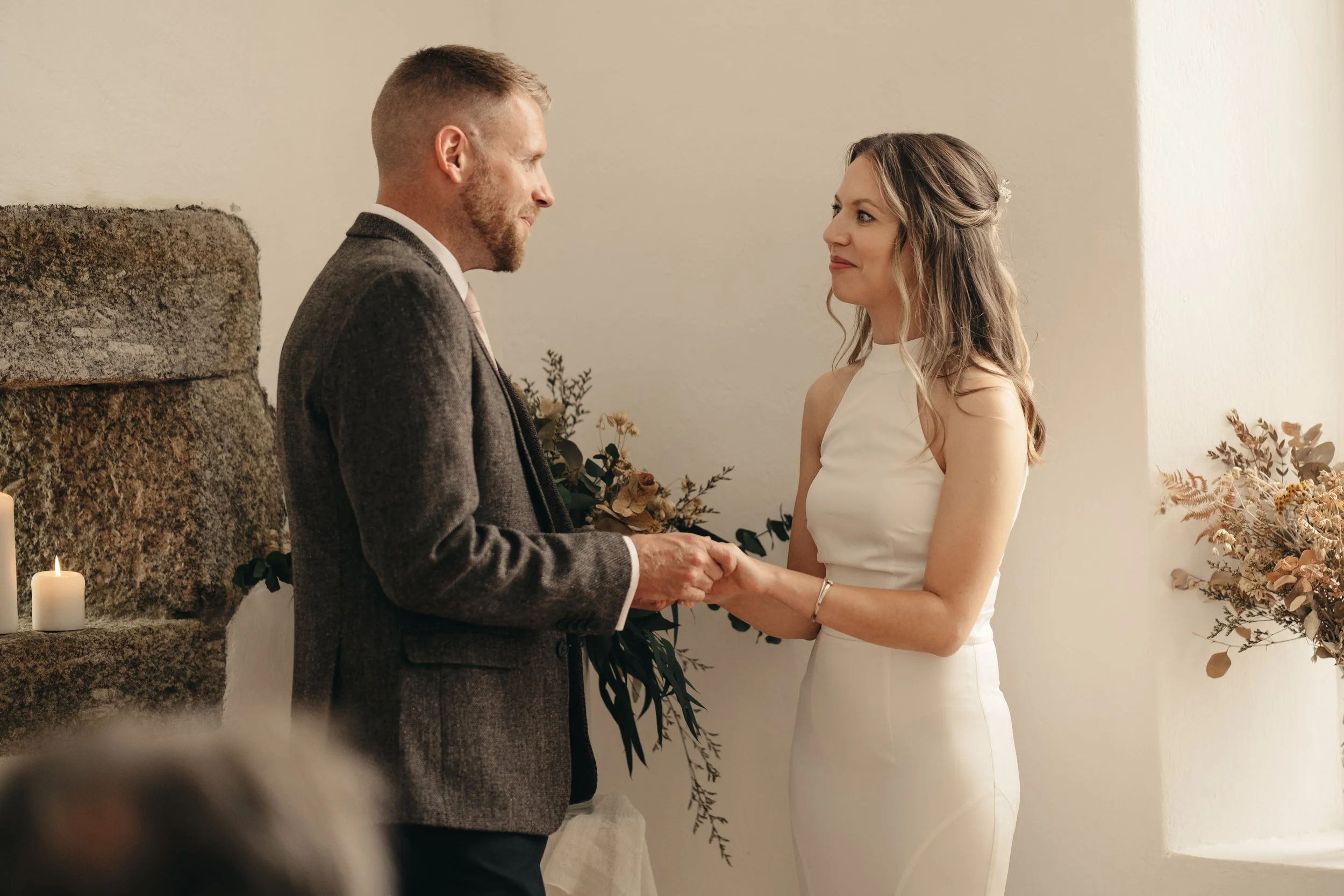 A couple exchanging vows during a wedding ceremony, with the groom in a grey suit and the bride in a white dress, holding hands and smiling at each other.  St Ives, The Block House elopement venue, Cornwall.