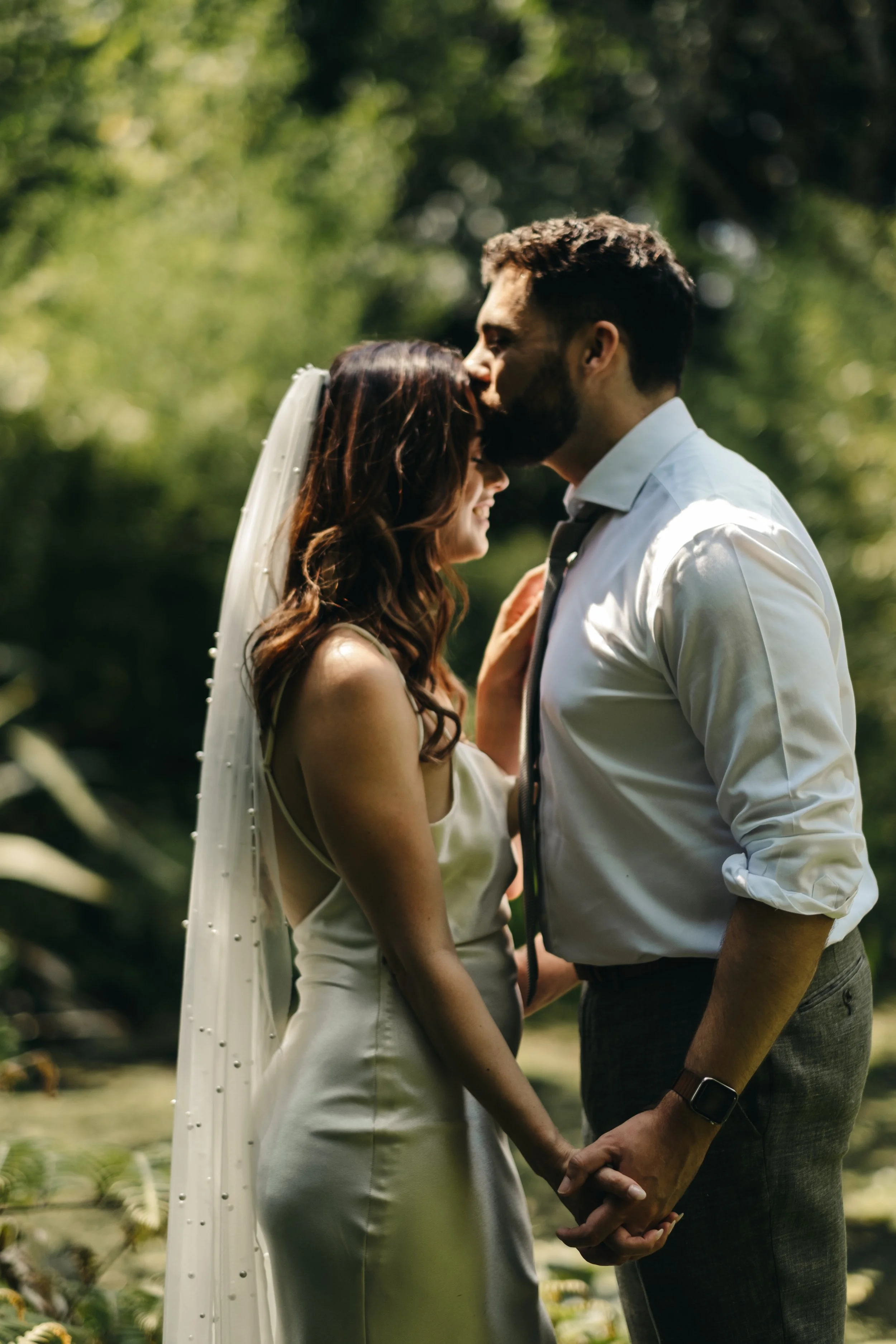 A bride and groom holding hands and sharing a kiss in a lush outdoor setting. Tremenheere Sculpture Gardens, Cornwall,  Cornwall Elopement Venue,  Cornwall Elopement Photographer.