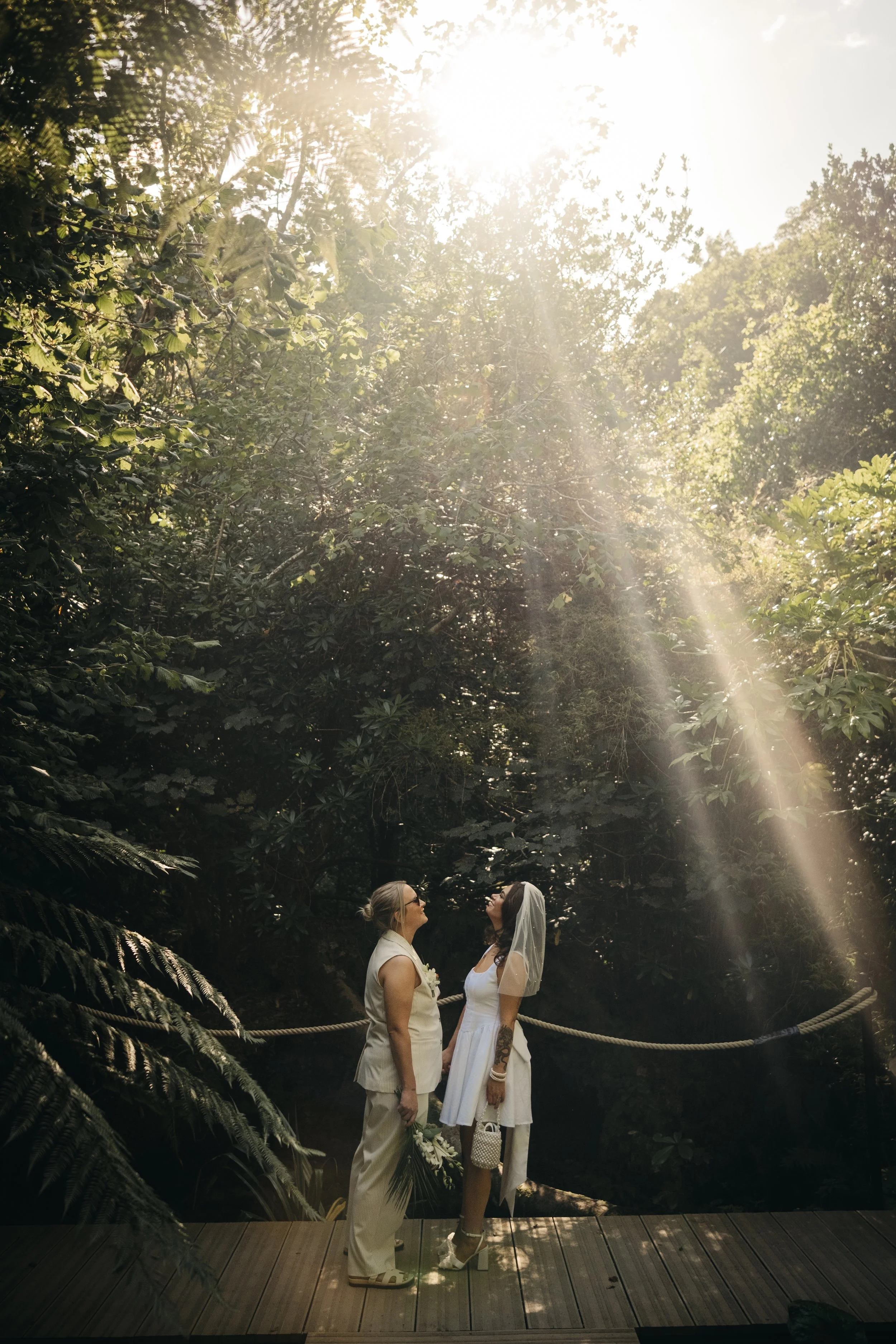 Two women, one in a white suit and the other in a white dress with a veil, standing on a wooden platform in a lush green forest with sunlight streaming through the trees. Tremenheere Sculpture Gardens, Cornwall,  Cornwall, Elopement Venue.