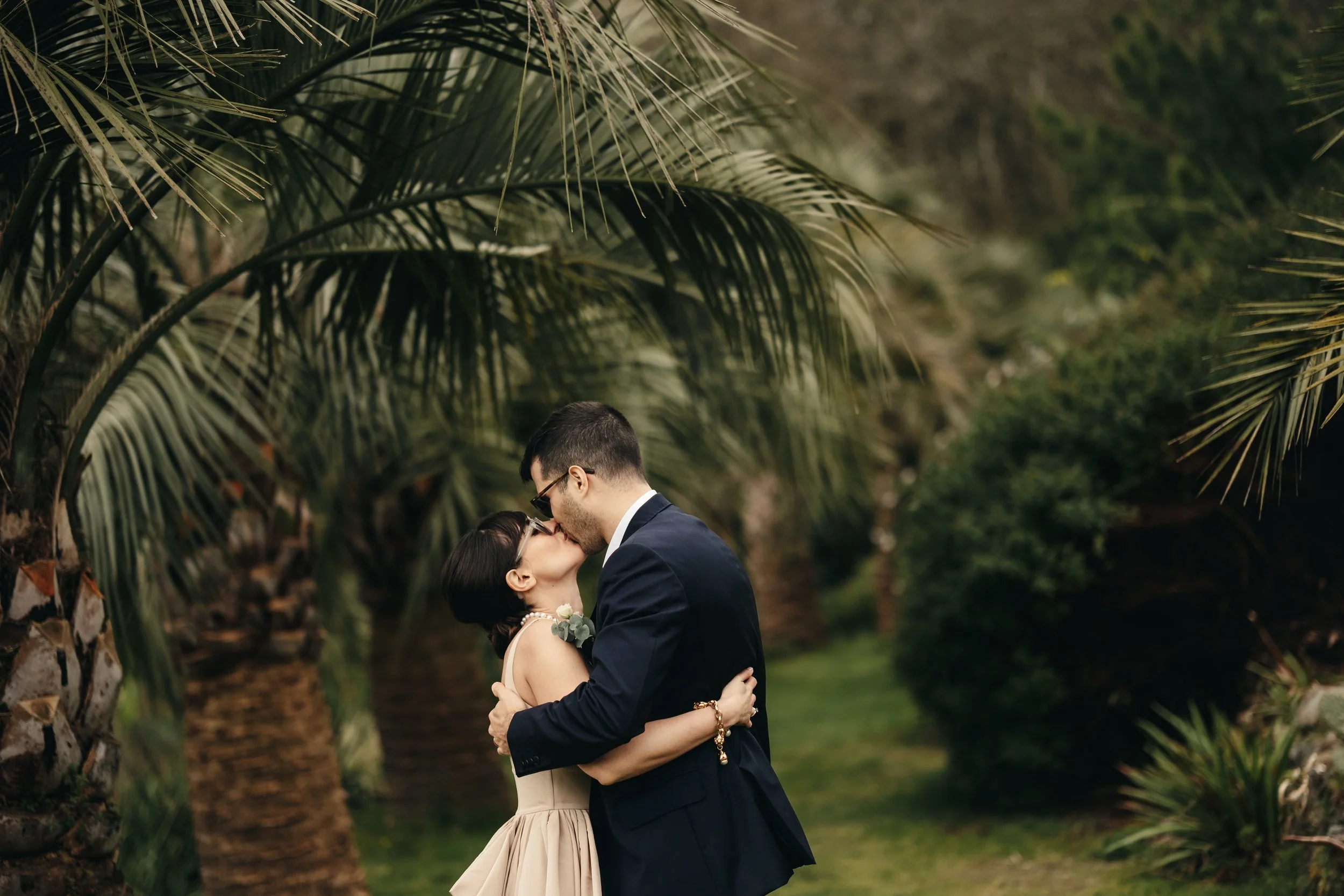 A couple sharing a kiss outdoors, surrounded by lush tropical plants and palm trees. Tremenheere Sculpture Gardens, Cornwall,  Cornwall Elopement Venue,  Cornwall Elopement Photographer.Tremenheere Sculpture Gardens, Cornwall,  Cornwall Elopement 