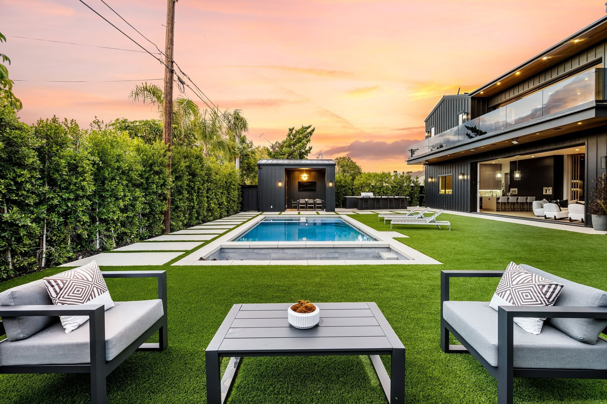 A modern backyard with a swimming pool, outdoor seating, and a view of a house at sunset. The yard has green artificial grass, and the house features large windows and a balcony. There are lounge chairs by the pool and a covered outdoor dining area.