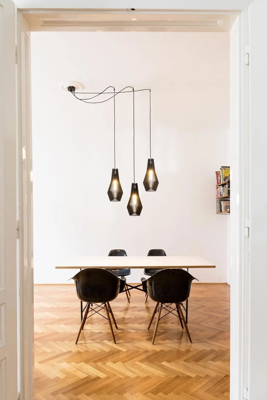 Das Bild zeigt drei handgefertigte Pendelleuchte SYMMETRY Version L über einem großen Esstisch von Egon Eiermann mit vintage Eames Plastic Armchairs DAR in einem großzügigen Wohnraum in einer Wiener Stilaltbauwohnung. Konfiguration I.