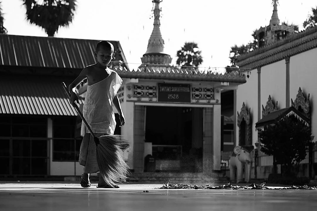 In the early morning light, a novice monk sweeps the temple courtyard while the stupa rises behind him. The dust catches the light as it lifts off the ground. It is the oldest kind of morning routine there is.