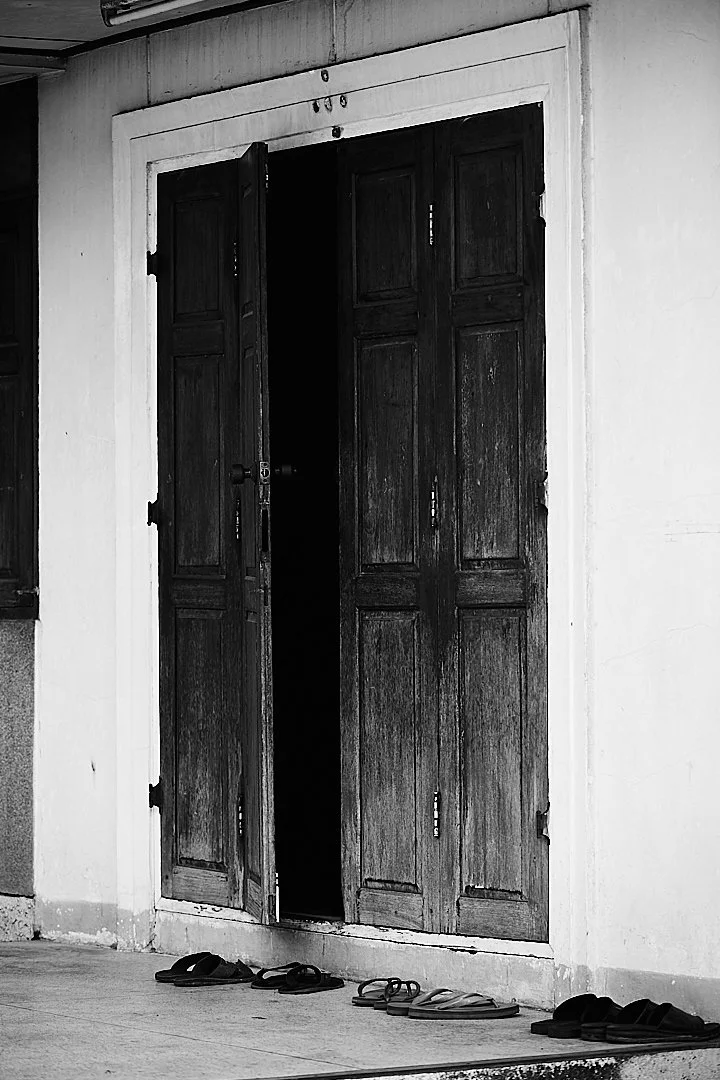 Three pairs of sandals outside a temple door. Someone is inside. The door is open just enough to let the dark in.