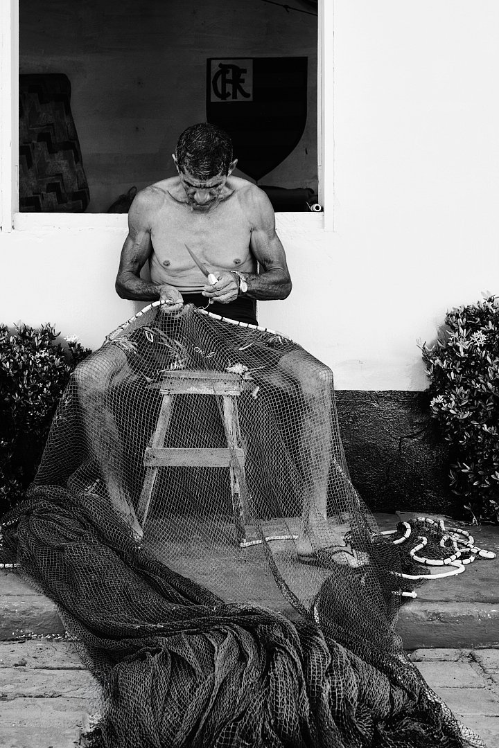 The nets never stay whole for long. He works through the damage methodically, needle moving through the mesh in a rhythm that needs no thought. The Flamengo crest on the wall behind him is the only color in the frame. Even here, football finds a way 