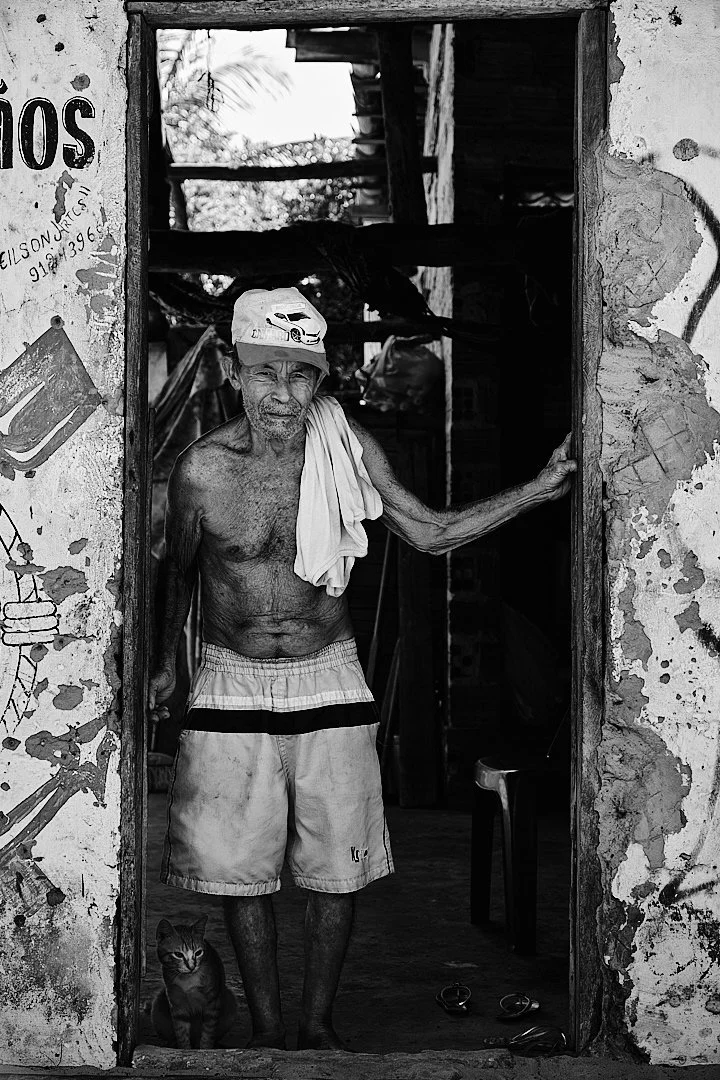 Daniel has lived in this doorway, or one very much like it, for most of his life. The cat at his feet is indifferent to the visitor. He is not. He stands with the ease of a man who has nothing left to prove, a towel over his shoulder, a small smile f