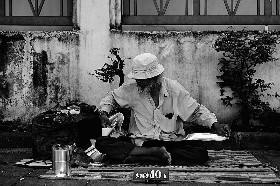 He sat against the wall with his things arranged around him and a small sign in front, 10 baht. The bonsai tree growing from the cracked wall behind him had been there longer than either of them.