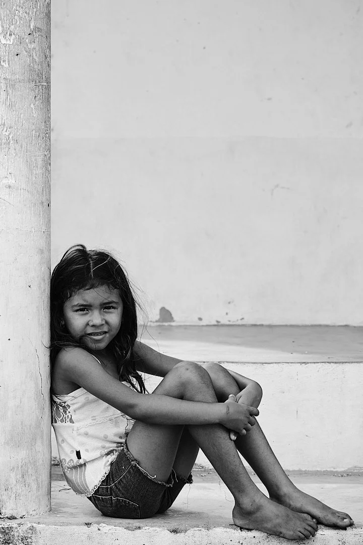 The children of Travosa grow up with the dunes at their back and the Atlantic at their feet. She leans against the worn plaster of a building older than she is, squinting into the light with the particular wariness of a child who has learned that str