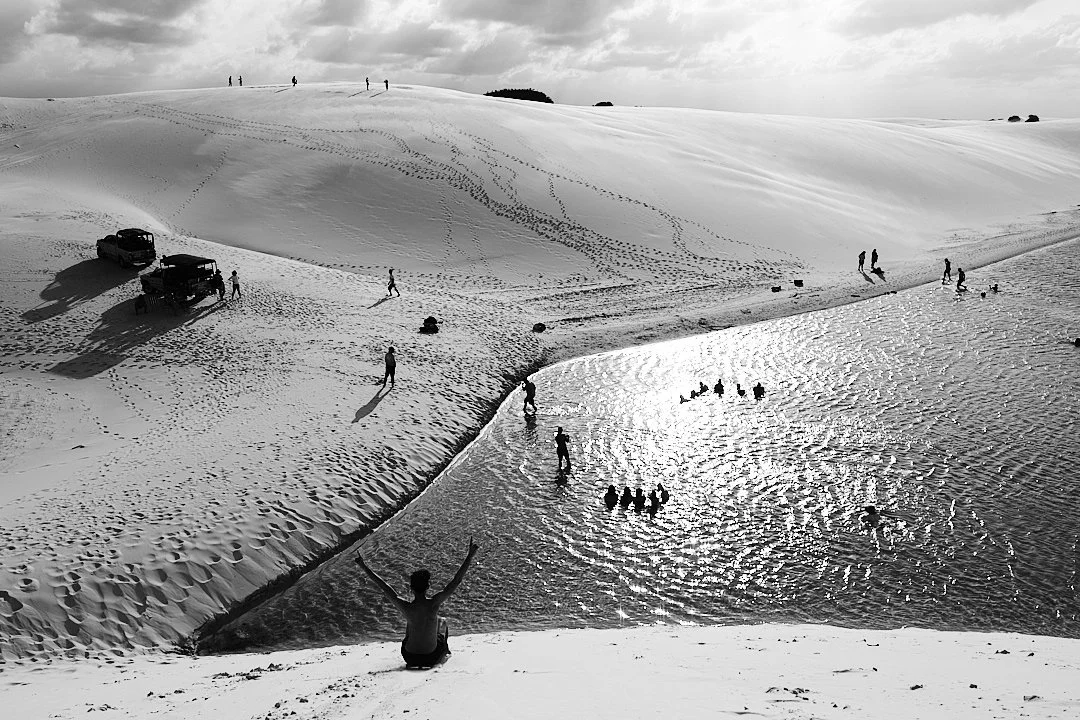 From above, the lagoon catches the sun and throws it back at the sky. Figures move through the water and across the sand in every direction. Jeeps sit at the edge where the sand hardens. The dunes go on past all of it. They were here before the villa