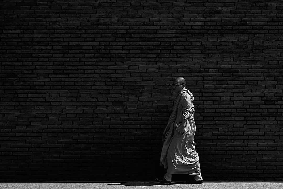 He moved along the wall at a pace the city could not touch. His shadow stretched out behind him on the pavement, longer than he was, going somewhere of its own.