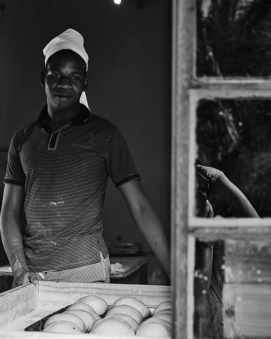 He came to Inhaca to be near family, not for the work itself. The bread sits ready in the foreground, waiting for the oven. He peers out the window at an island that is still becoming home.