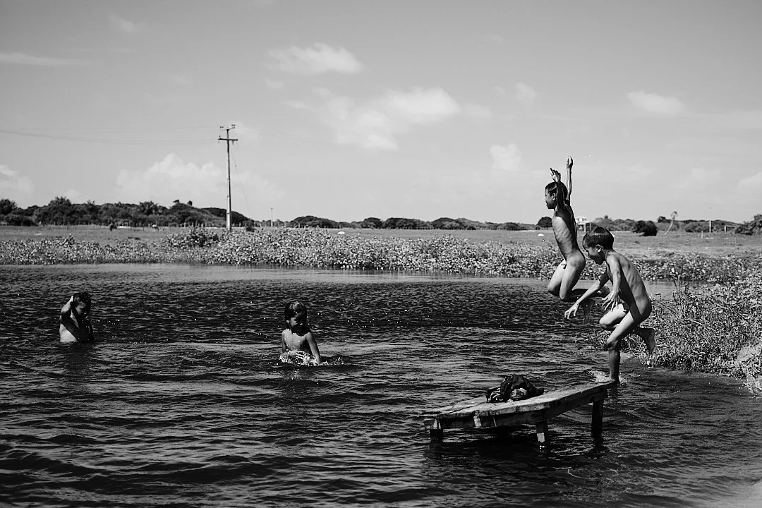 When the heat comes down the children go to the water. A plank of wood, a shallow lagoon, and the kind of afternoon that asks nothing of you. One boy launches himself into the air with his whole body. The others wait their turn. This is the other sid