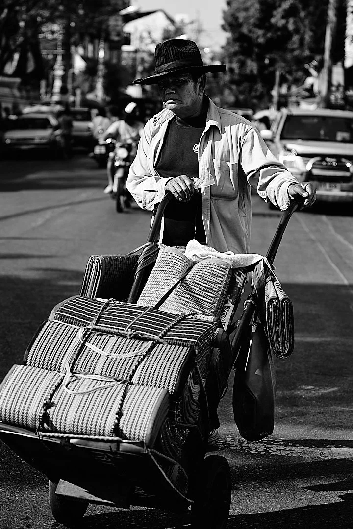 He pushed his load through the midday traffic without hurrying, hat pulled down against the sun, eyes forward. The city moved around him the way water moves around stone.