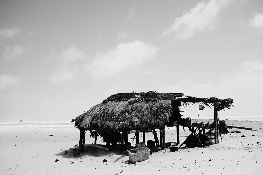Not everything lasts. On the beach, a palm frond shelter leans into the wind, holding its gear and its nets and its ropes underneath like a man hunching against the cold. No one is here right now. Someone will be back.
