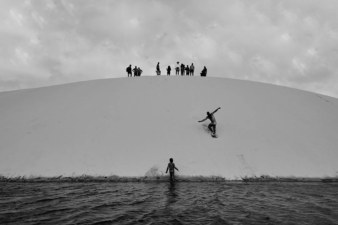 On weekends the dunes draw people from outside the village. A young man carves down the face of a dune on a board while a crowd watches from the ridge above. A child wades in the water at the base. The dunes belong to everyone but they started here, 