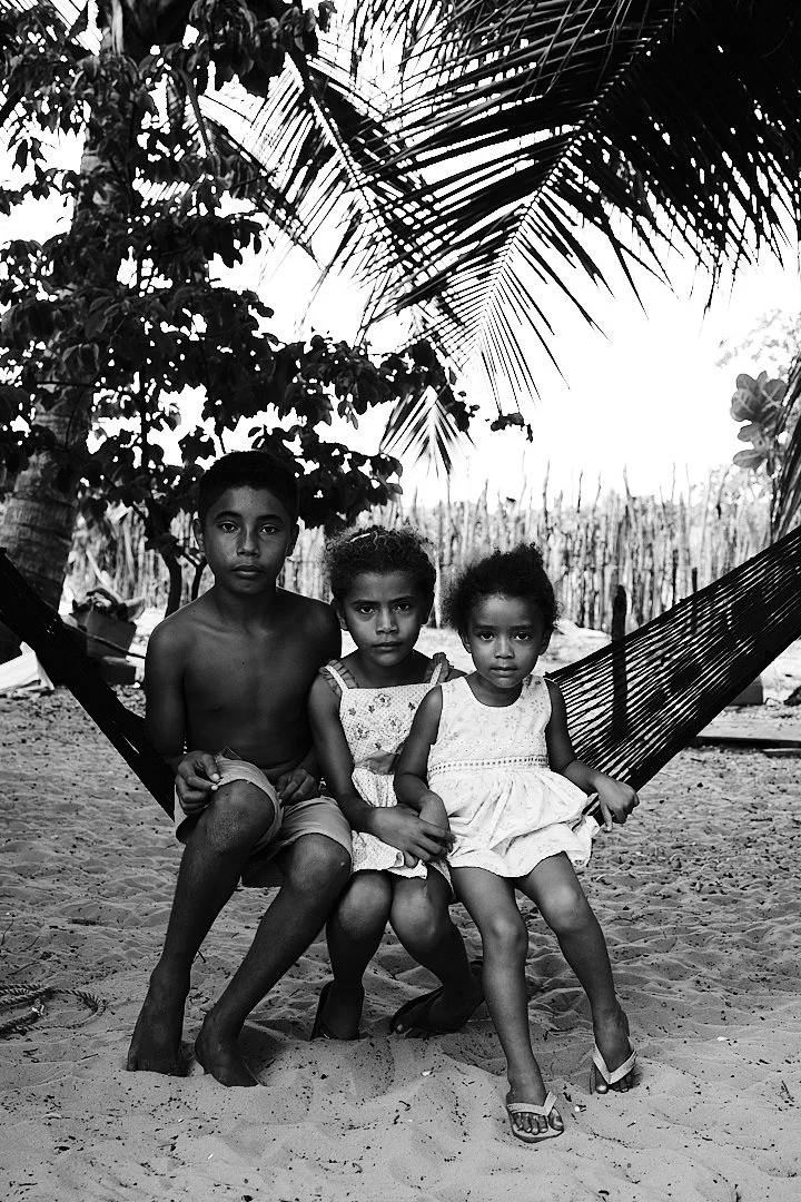 Three siblings sit together in a hammock strung between the palms, the oldest holding the younger two in place. They look at the camera without smiling, without performing. Just looking. There is a seriousness in their faces that has nothing to do wi