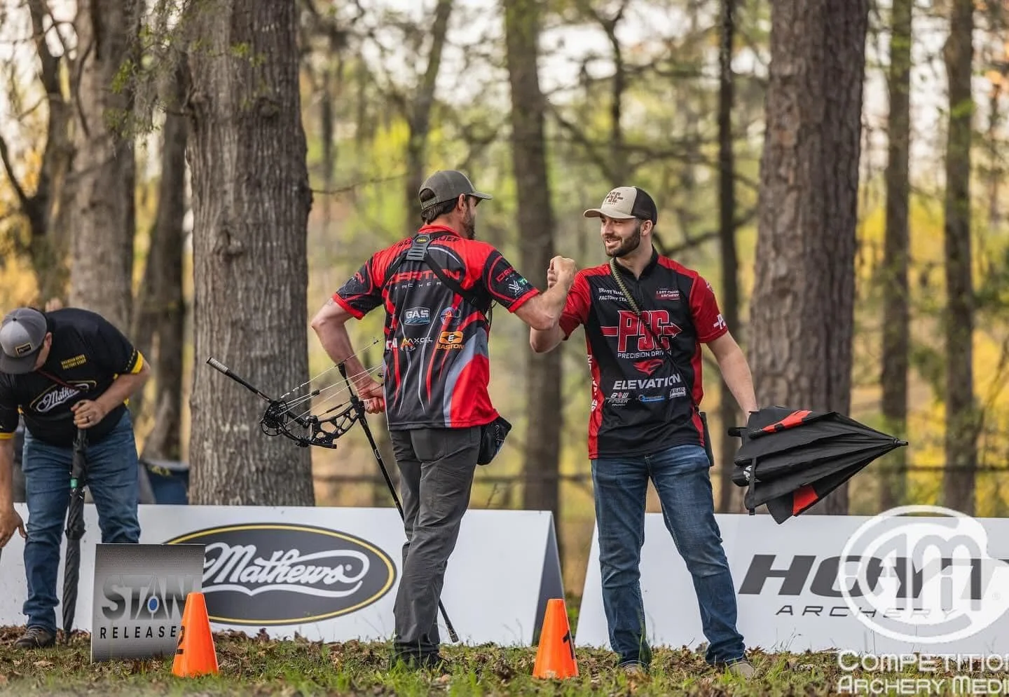 Two men in archery jerseys exchanging a fist bump in a wooded outdoor area during a competition, with one holding a bow and a folded umbrella nearby.