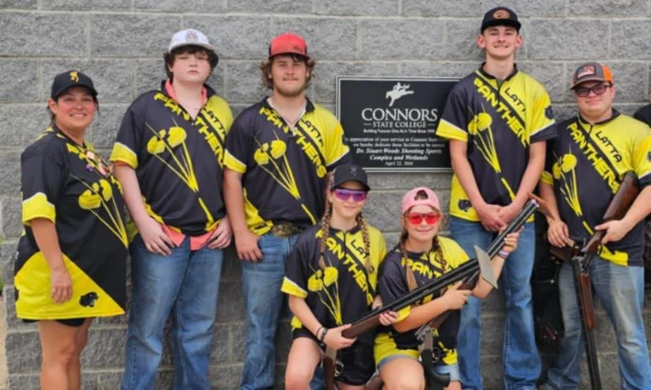 A group of young archers in matching yellow and black jerseys pose in front of a gray brick wall with a sign for Connors State College. They are holding bows and arrows, wearing sunglasses and hats.