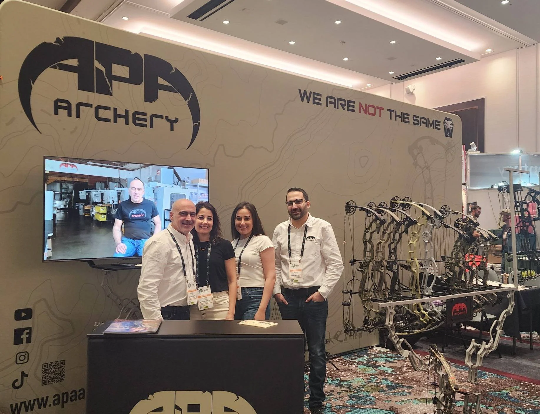 Group of four people standing at an archery trade show booth with a large bow sculpture, a TV screen displaying a man, and branding for APA Archery, with camouflage-patterned carpet.