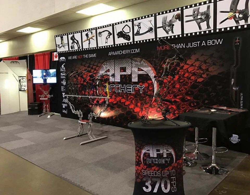 Archery exhibition booth with a large black and red backdrop displaying the AP archery logo, a bow and arrow graphic, and the slogan 'More than just a bow.' The booth features a metal bow sculpture, a tall table with AP archery branding, a monitor on
