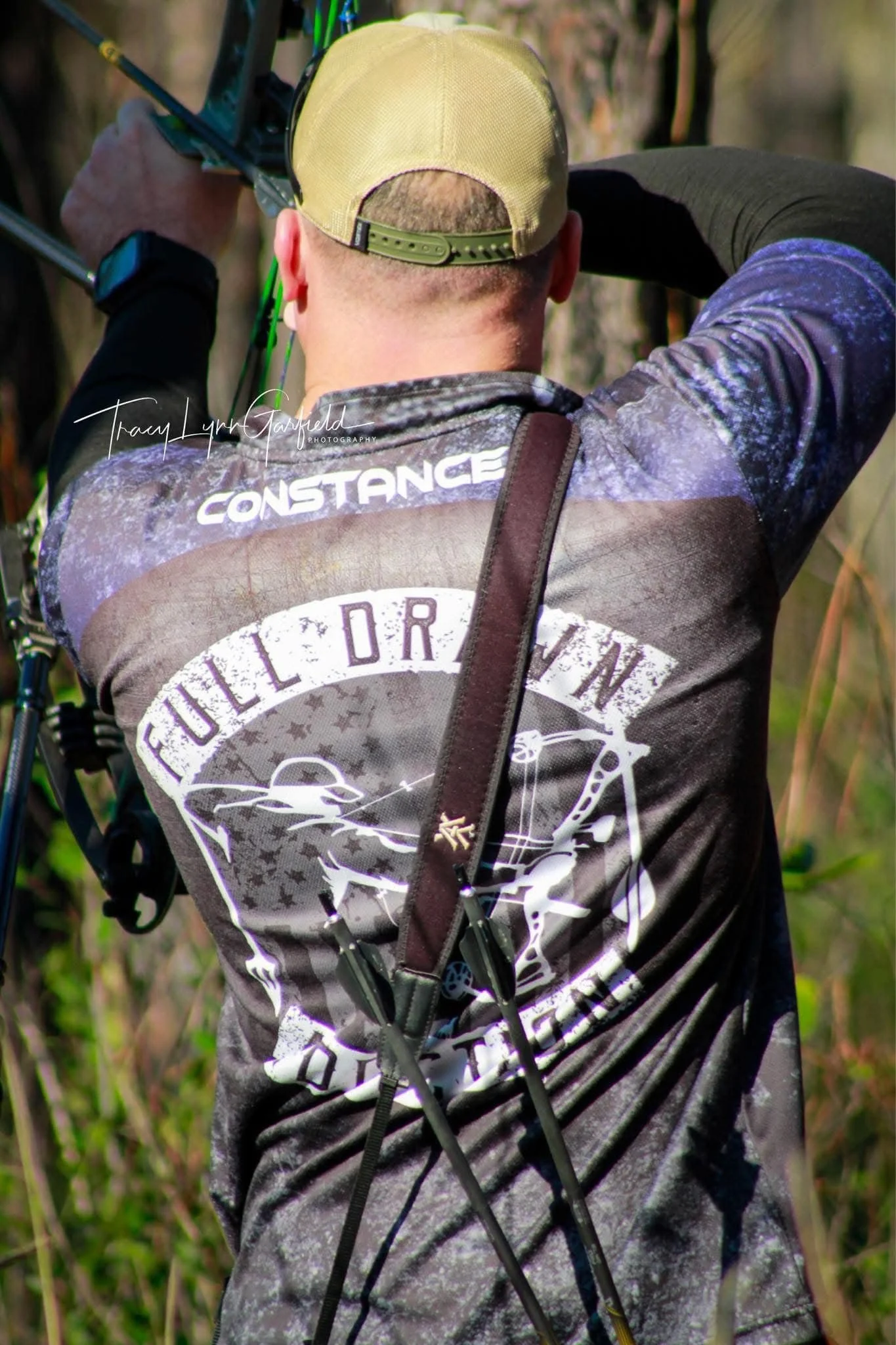 Back of a person wearing a camouflage shirt and a yellow cap, carrying a bow, in a forest environment during daytime.