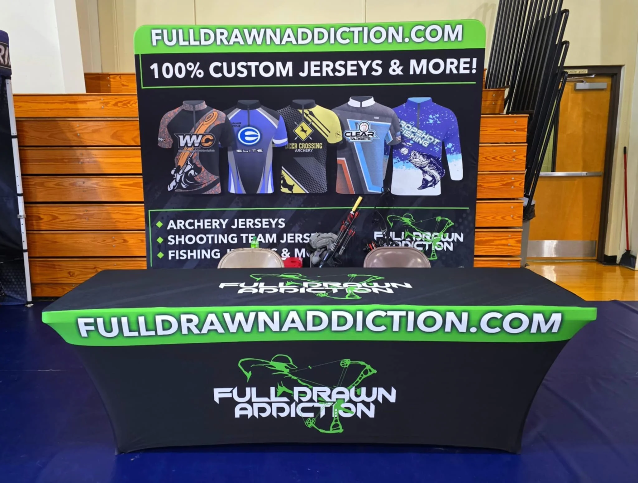 A promotional booth featuring a large sign advertising custom jerseys and more at fulldrawnaddiction.com. The sign depicts various jerseys including archery, shooting team, and fishing. The table in front has a black cloth with green trim displaying