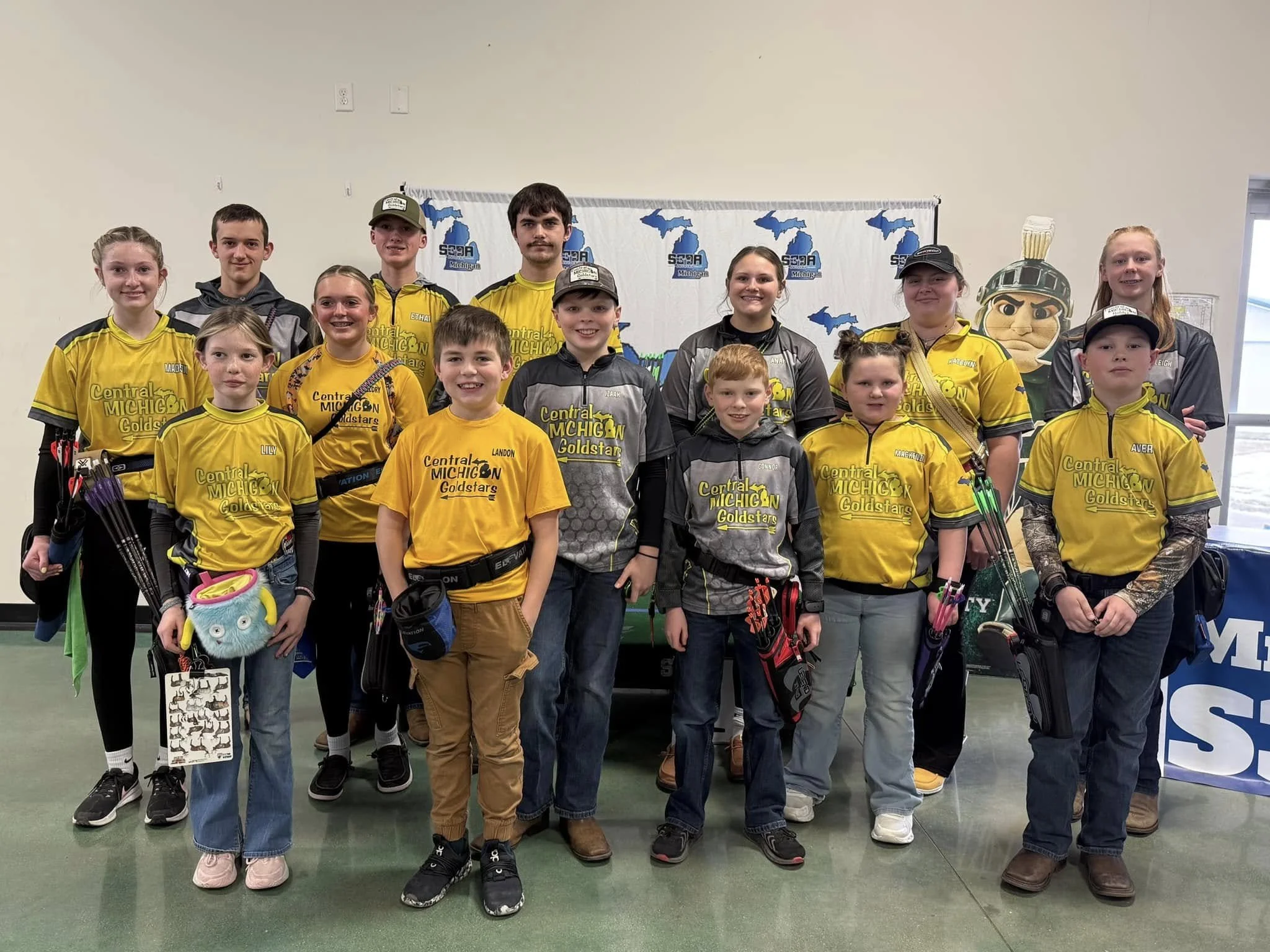 A group of children and teenagers in yellow and gray archery uniforms posing for a photo indoors at a competition or event.