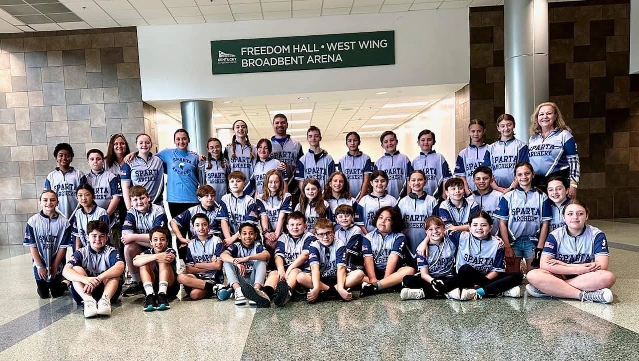 A group of children and adults in matching blue and white sports jerseys, posing for a photo in front of a sign that reads 'Freedom Hall West Wing Broadbent Arena' inside a building.