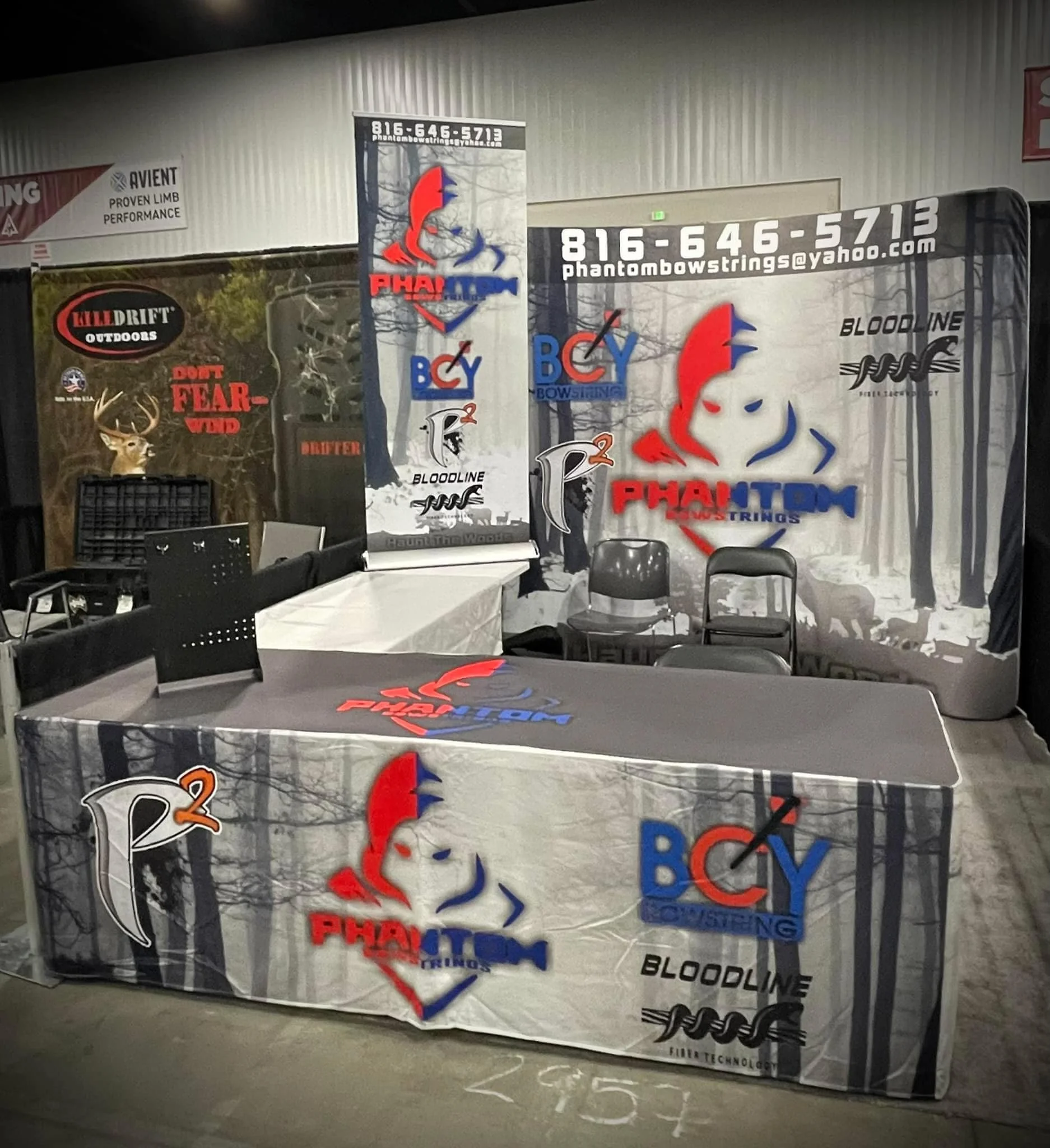 Booth display for Phantom Bloodline Bowstrings featuring banners and table cloth with logos and contact information, set in an indoor event space.