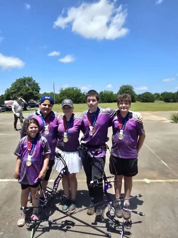 Group of five children in purple sports jerseys with medals, standing outdoors with archery equipment, smiling at the camera on a sunny day.