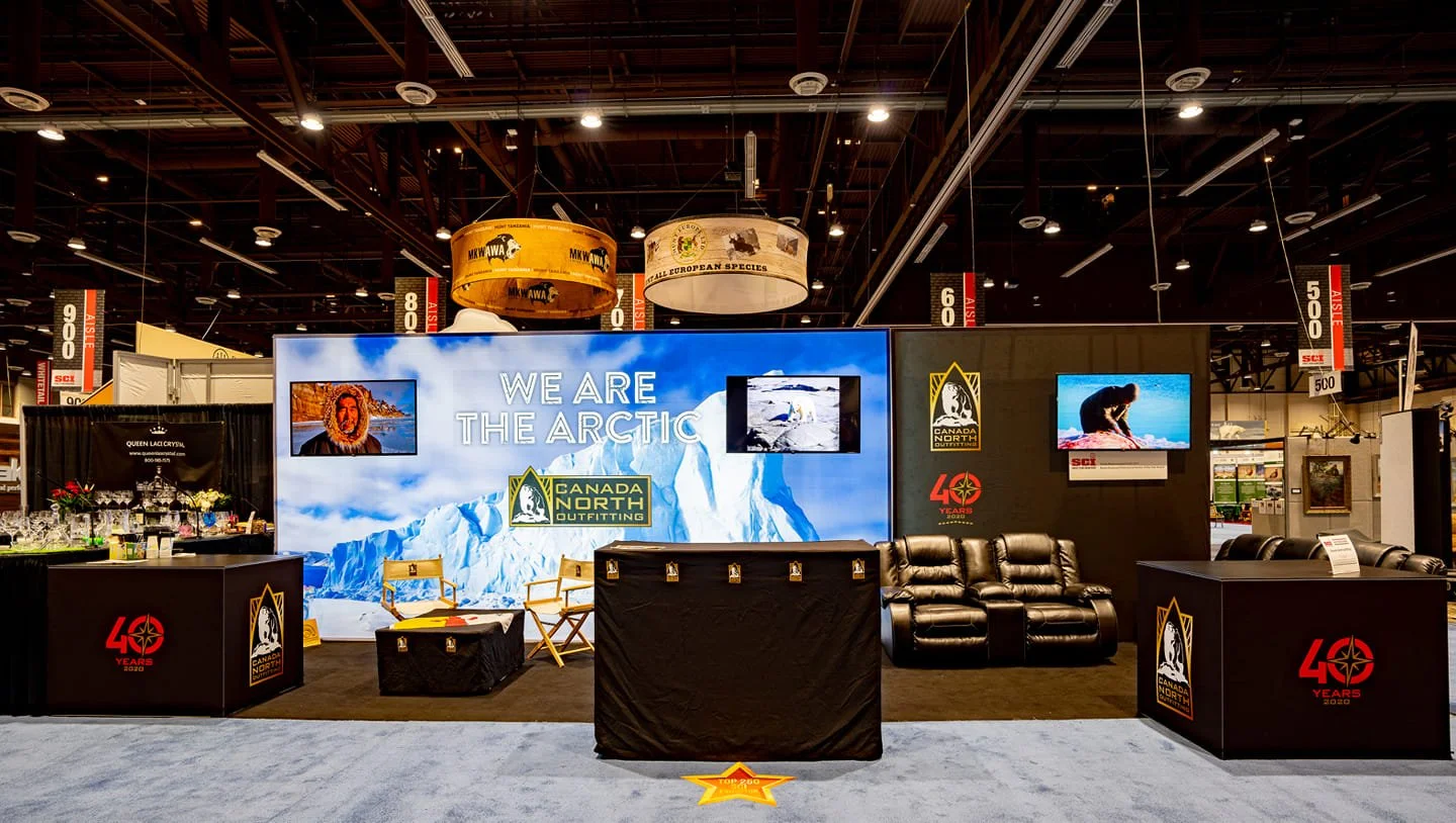 Booth at a trade show with large digital screens displaying Arctic imagery, with text 'We Are The Arctic' and logos of Canada North Outfitting and 40 Years. There are chairs, a black leather couch, and display counters with promotional materials.
