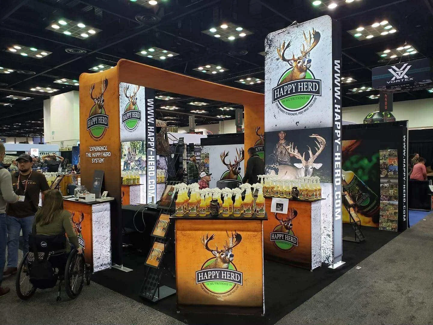 Trade show booth for Happy Herd Nutrition featuring images of a buck and antlers, with product bottles displayed on counters and promotional posters, at an indoor convention center.