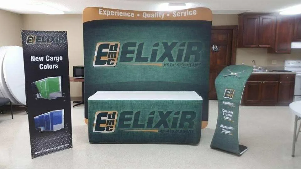 Trade show display for Elixir Metals Company featuring logos, a table with branded cover, and side banners advertising cargo colors, roofing, custom frame parts, and aluminum siding in a room with kitchen cabinetry.