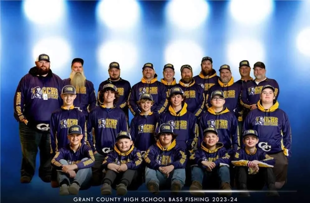 A group of young students and adult coaches from Grant County High School Bass Fishing team posing in matching navy blue and yellow jackets and hats with the school's logo, set against a blue background with bokeh lights, for the 2023-24 season.