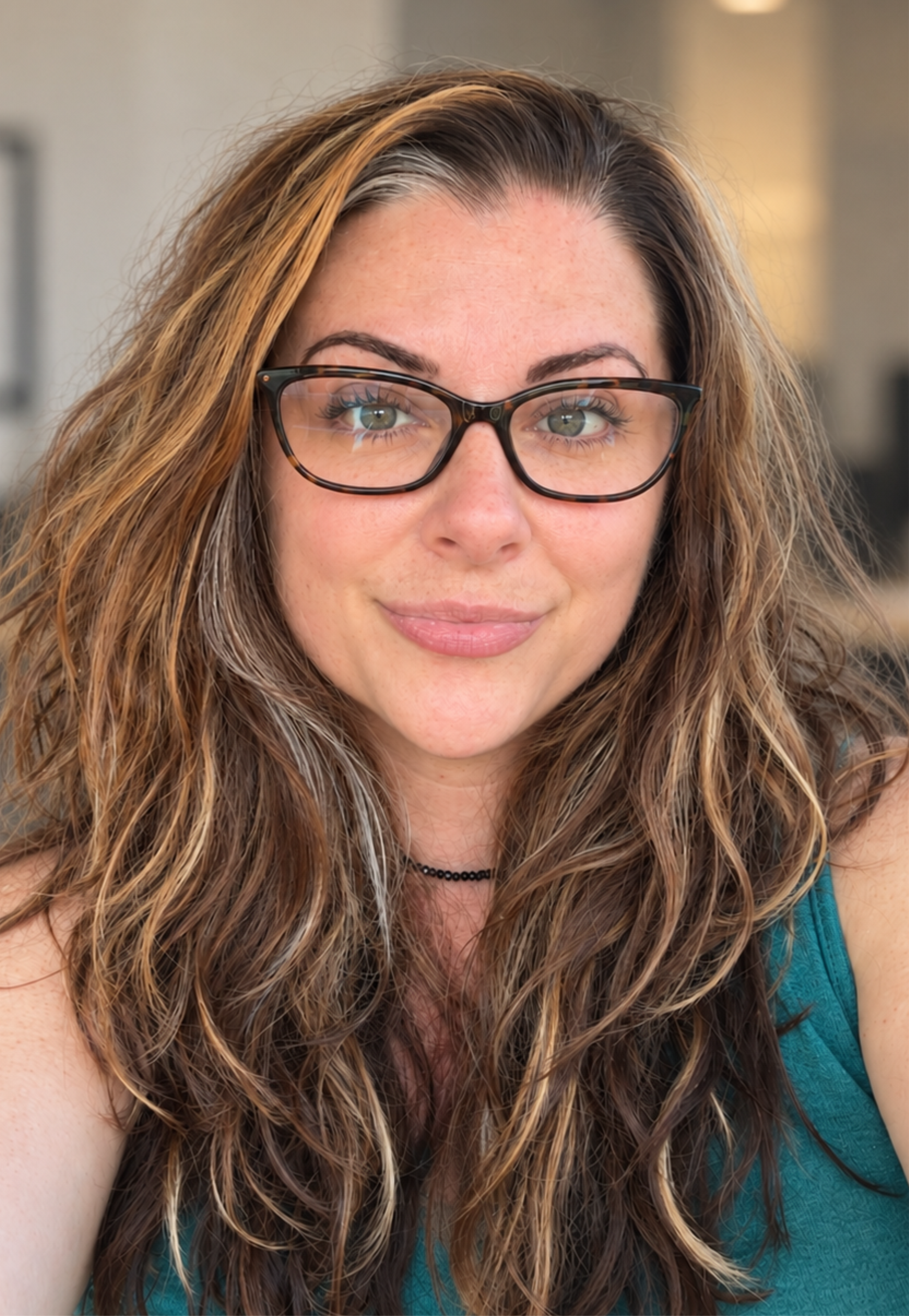 Close-up of a woman with long, curly brown hair wearing glasses and a teal top, smiling softly.