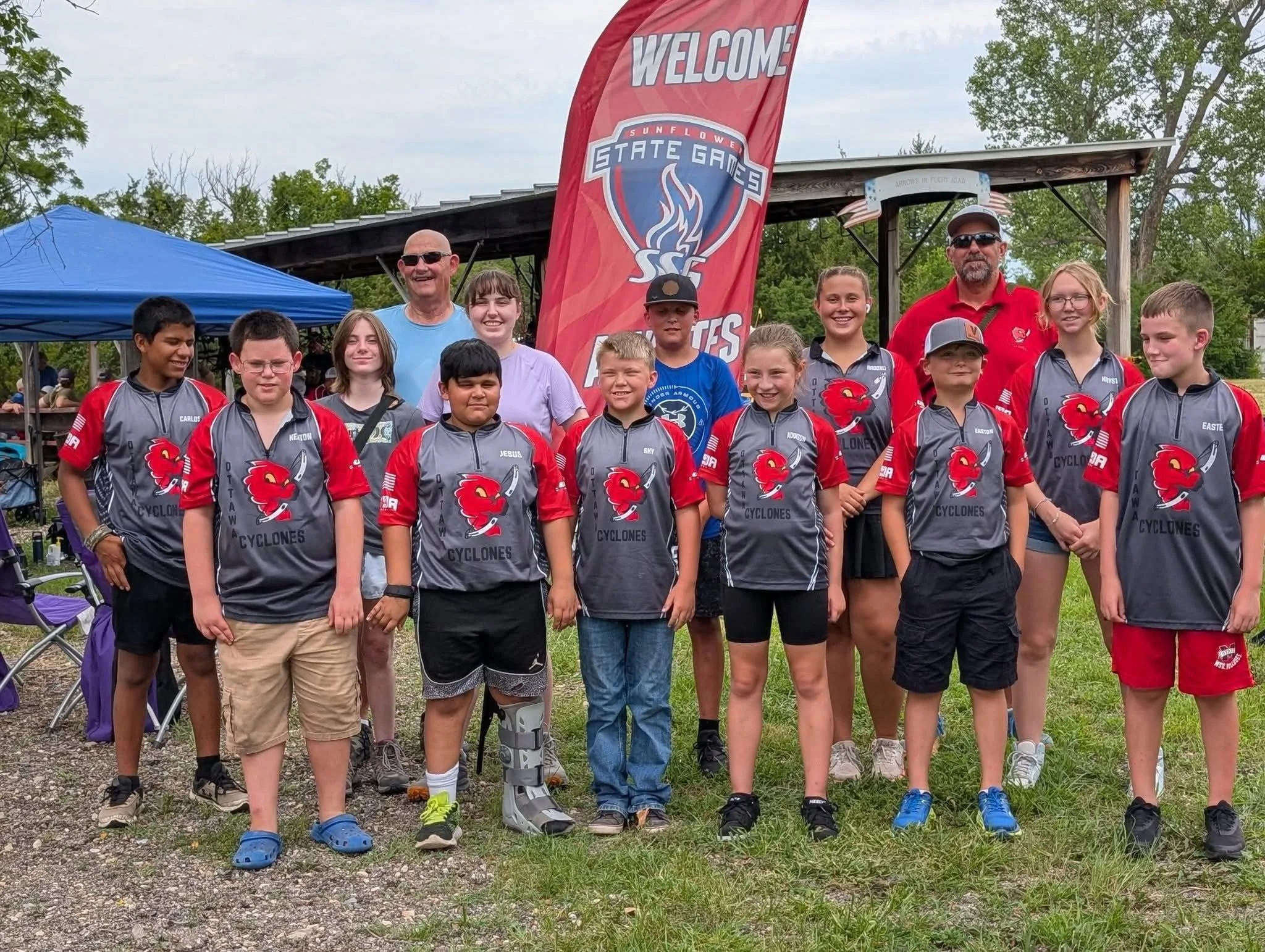 Group of young athletes in matching team jerseys, with two adults behind them, standing outdoors in front of a red banner that reads 'Welcome' and 'Sunflower State Games'.