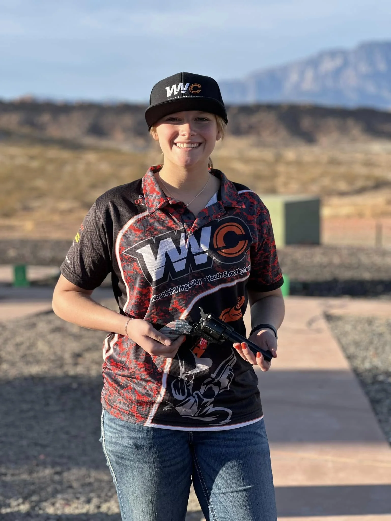 A young woman outdoors wearing a black WAC baseball cap and a matching sports jersey, smiling and holding a gun, with a mountainous landscape in the background.