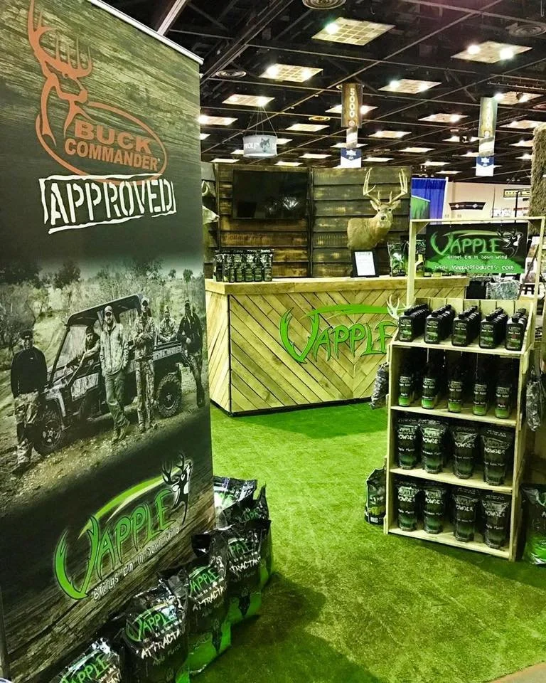 Display booth for Buck Commander and Vapple products featuring deer mounts, product shelves, and promotional banners, with artificial grass flooring.