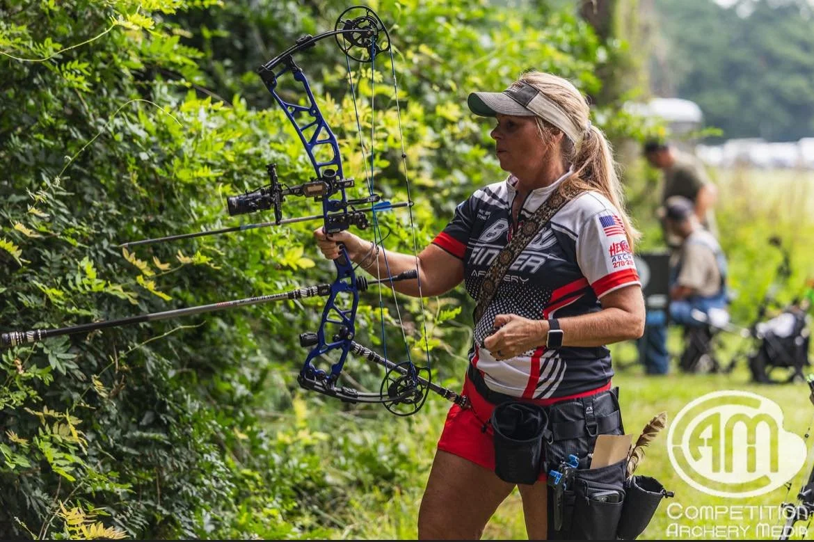 A woman in sportswear practicing archery outdoors, aiming her compound bow at a target amidst green foliage. She has a visor and a quiver attached to her waist, with other competitors visible in the background.