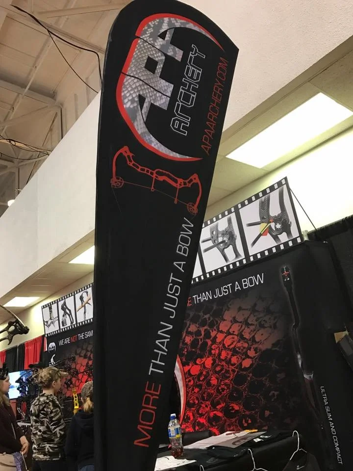 A black vertical flag advertising archery equipment with the words "More Than Just a Bow" and an outline of a bow. The background includes a booth setup at an indoor event.