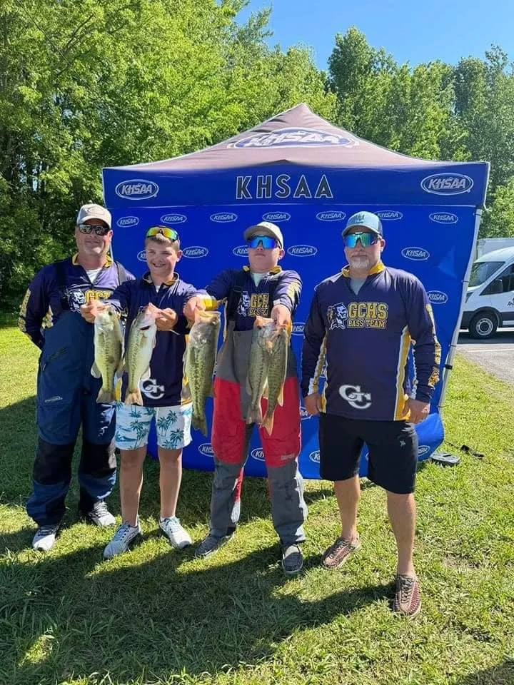 Four men standing outdoors in front of a blue tent with 'KHSAA' branding, holding large fish. They are dressed in fishing gear with 'GCHS' and 'Bass Team' logos, indicating a fishing competition or team event.