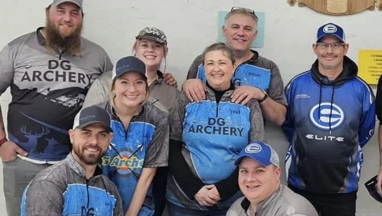 Group of seven people smiling, wearing archery gear, indoors with a plain wall background.